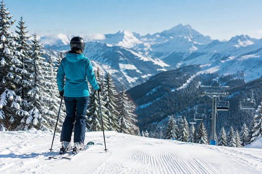 Skieur de dos avec casque et masque, arrêté au bord d'une piste damée dans les Alpes françaises, sapins enneigés et sommets au soleil