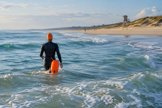 Nageuse de dos en combinaison et bonnet vif, bouée de natation orange attachée à la taille, entrant dans l'océan sur une plage atlantique